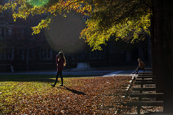Two students outside on an autumn day, one standing, one seated.