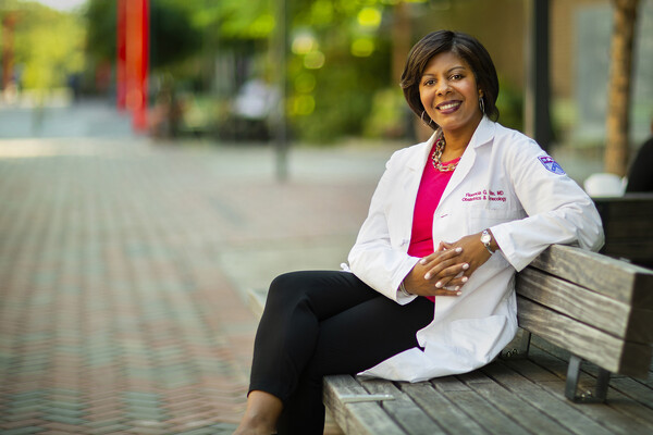 Person in a lab coat sitting on a wooden bench outside. 