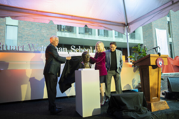 Two people pulling cloth off of bronze bust next to a podium with third person looking on
