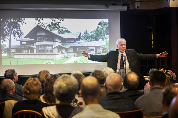 Paul Hendrickson speaking at a podium in front of a seated crowd, his arms outstretched, with a photo of a house on the screen behind him.