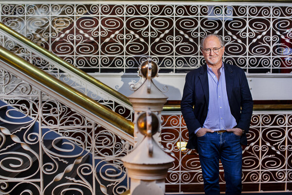 A person in jeans, a blue button down shirt and a blazer stands with hands in pockets in front of an ornate white-iron stairwell.