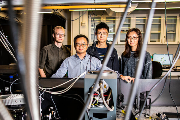 four people standing in front of an optics table behind a tangle of wires in a lab in 