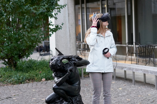 Person wearing a virtual reality headset looks to the side, standing next to a statue facing the same direction