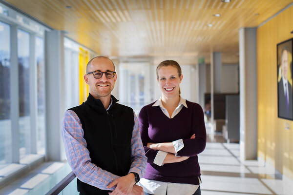 Two people standing in a building entryway. Windows are on their left, a yellow wall with a portrait are on their right. They're both looking into the camera and smiling.