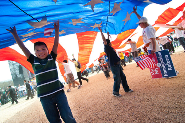 Several adolescents stand under a giant American flag like a parachute aloft at an immigration reform rally.