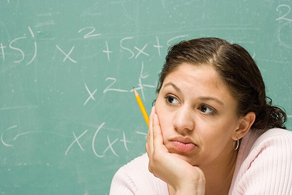 person looks perplexed holding a pencil staring into middle distancw with chalkboard behind them with math equations.