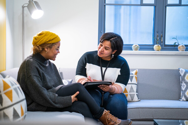 Two people sitting on an L-shaped couch amidst four pillows. The one on the right is holding a clipboard.