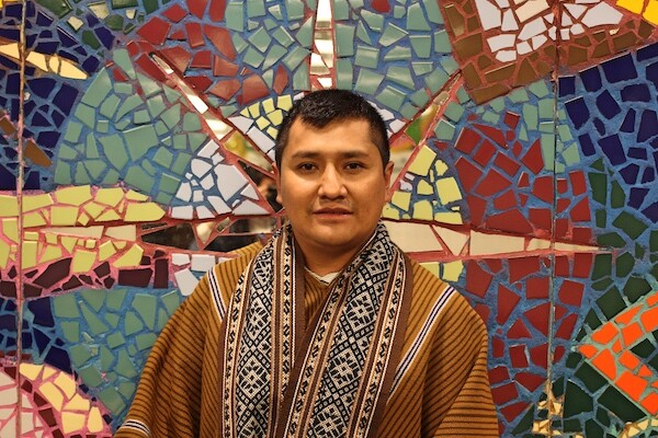 Man wearing traditional Andean clothing stands in front of mosaic