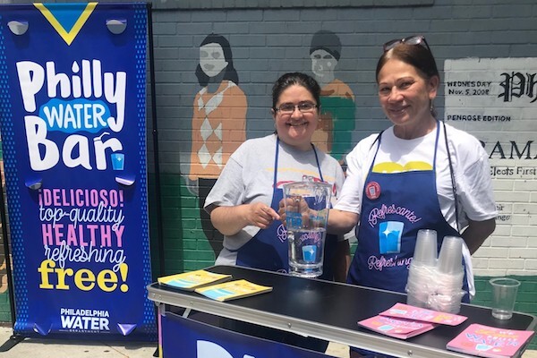 Two smiling people with a pitcher of water stand next to a sign saying "Philly Water Bar"