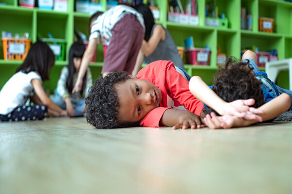 a young kindergartner lays on the ground looking unenthused while other kids play in the background