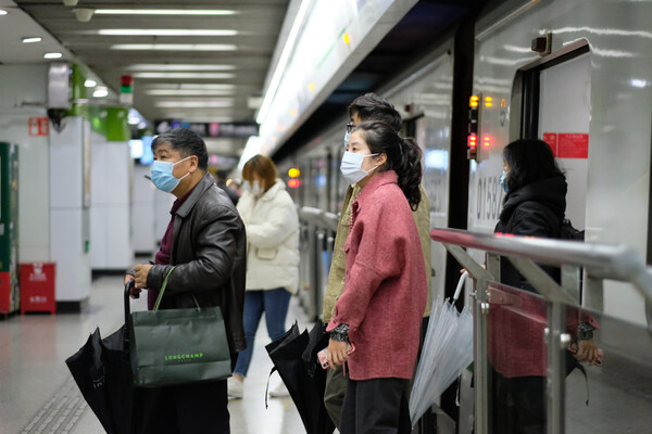 People in a subway station in Shanghai wearing surgical masks. One is carrying two bags, another is carrying a phone, a third is holding an umbrella.
