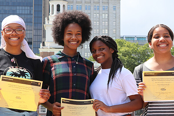Four students hold certificates from Philadelphia Writing Project smiling and standing outdoors