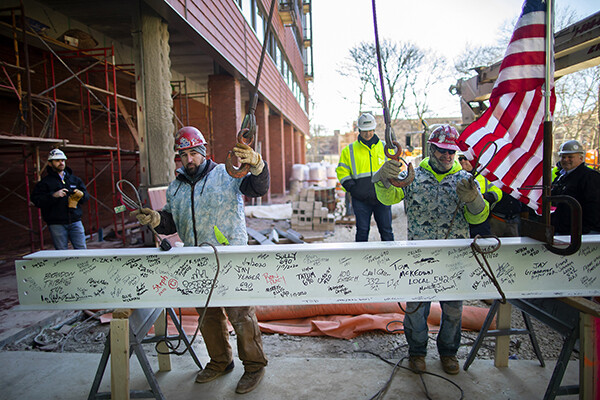 Two construction workers attach a crane to wires around a beam covered in signatures at the topping-off ceremony for New College House West
