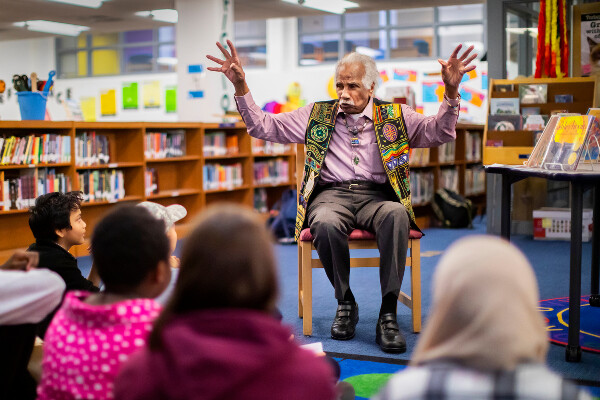 Ashley Bryan gestures with his hands open at a library at an elementary school reading his books to children seated on the ground.