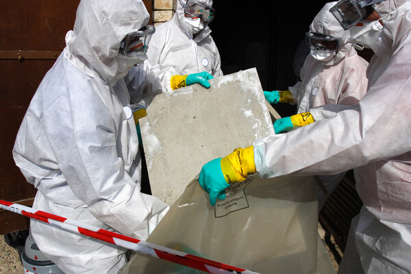Four workers wear protecting clothing while placing a board with asbestos into a plastic bag.