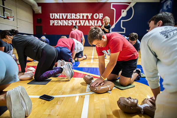 a student performs chest compressions on a mannequin on the floor of a gymnasium, large University of Pennsylvania banner hangs on the far wall in the background.
