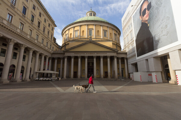 A person with two dogs walking in front of an empty plaza in front of the Cathedral of San Carlo al Corso in Milan, Italy
