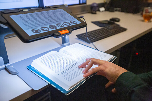 A hand on an open book attached to an assisted reading computer at the Weingarten assisted learning center