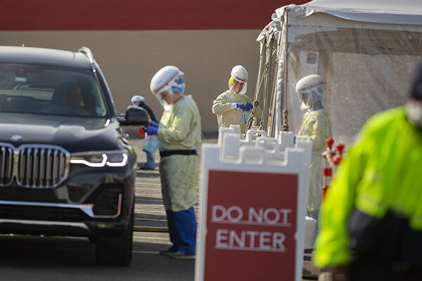 Three medical personnel outside in protective gear working at a drive-through COVID-19 testing site, a Don Not Enter sign in front of the vehicle