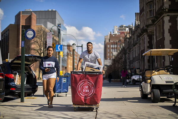 Two Penn students walk down a sidewalk on campus, one pushes a move-out cart.