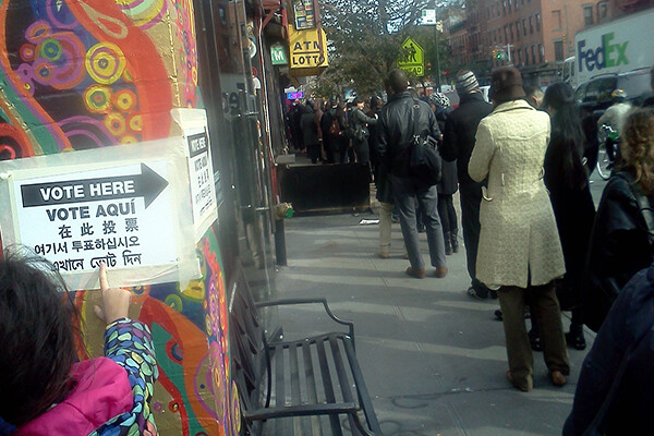 A line of people waiting to vote outside on a city sidewalk, a child points to a Vote Here sign taped to a colorful wall painted with a mural.