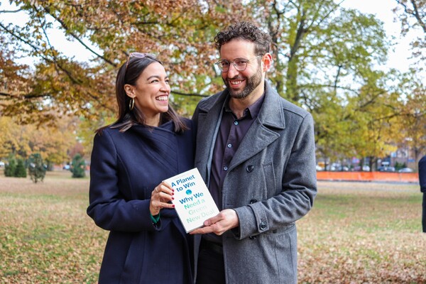 Two people standing outside on a lawn covered in leaves, holding a book titled "A Planet to Win, Why we need a Green New Deal." 