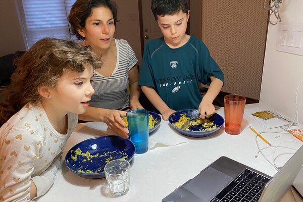 A professor and two children eating from bowls at a kitchen counter with a laptop computer open in front of them. 