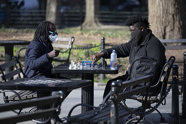 Two people sit in a public park in a city playing chess at a park table wearing protective face masks.