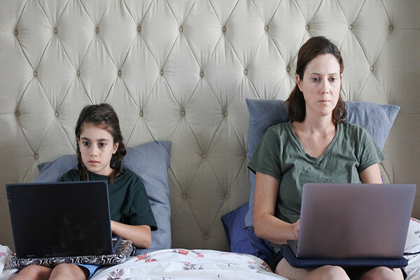 A parent and young child sit upright next to each other on a bed with laptops in their laps