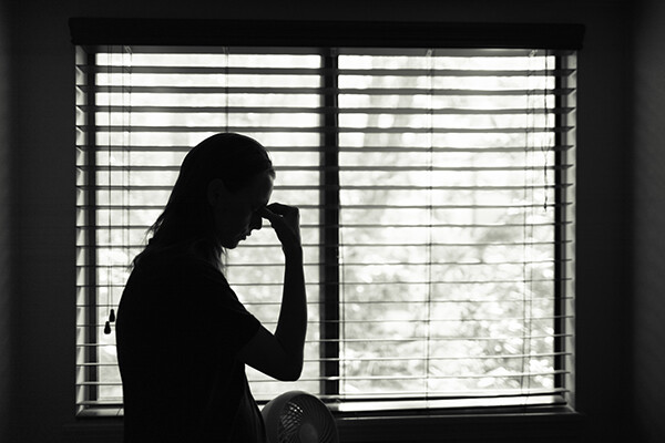 Black and white image of a person standing in front of an open blind, a hand held to the forehead in consternation.