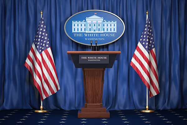 Empty podium with seal of the White House flanked by American flags with plaque reading The White House hung from curtains in background