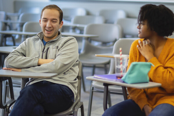 Two people sit at school desks, person on the left is smiling with arms cross as person on the right looks toward him.