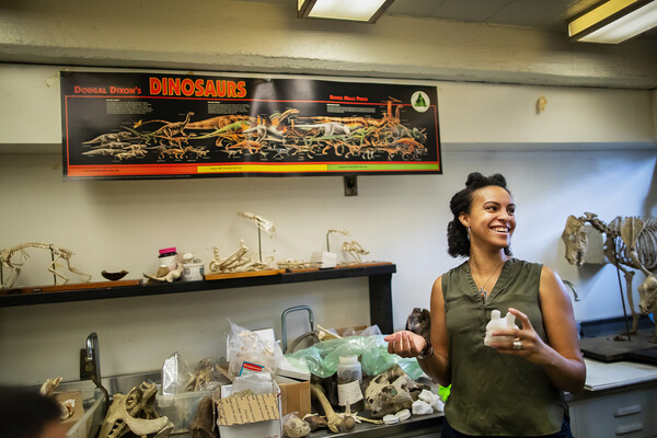 A person standing in front of a lab bench full of bones. On the wall hangs a poster that reads: "Dougal Dixon's Dinosaurs."