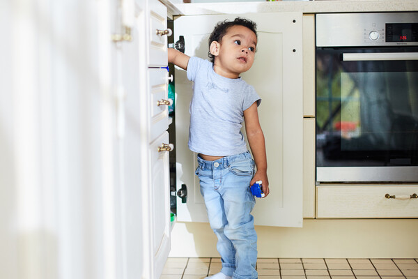 Child standing with a hand in an open cabinet in what appears to be a kitchen. A clock on the stove nearby reads 3:26.