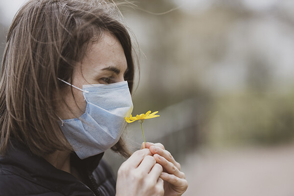 Person wearing a protective face covering holds a flower to their nose in attempt to smell its scent.