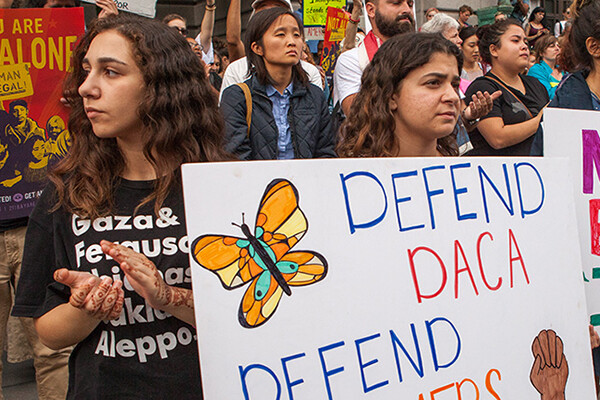 crowd of people at a demonstration holding signs, one reads DEFEND DACA