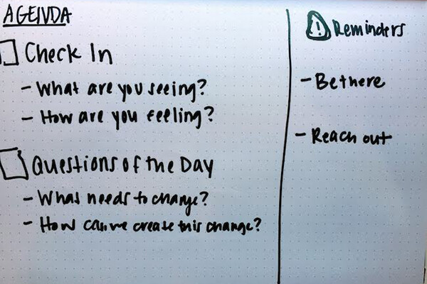 Writing on a whiteboard reading: Check-in, what are you seeing? What are you feeling? Question of the day: What needs to change? How can we create this change? Reminders: Be there. Reach out.