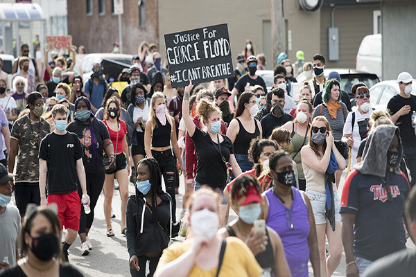 A crowd of people wearing masks march in the streets, one protester holds a sign reading "Justice for George Floyd."
