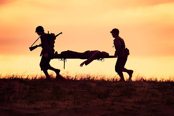 Two soldiers on a field carry a stretcher with a wounded soldier.