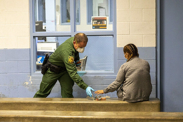 Uniformed border patrol personnel at a border processing center in a face mask and latex gloves hands an item to a detained person sitting on a bench.