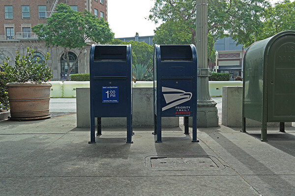 Two U.S. postal service mail boxes sit side by side on a sidewalk with trees behind them and a the first few floors of a red brick building on the left in the background