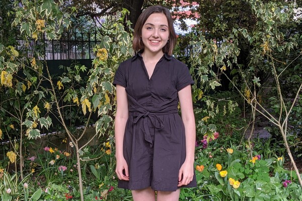 Person standing outside in front of trees and flowers hiding an iron fence. 