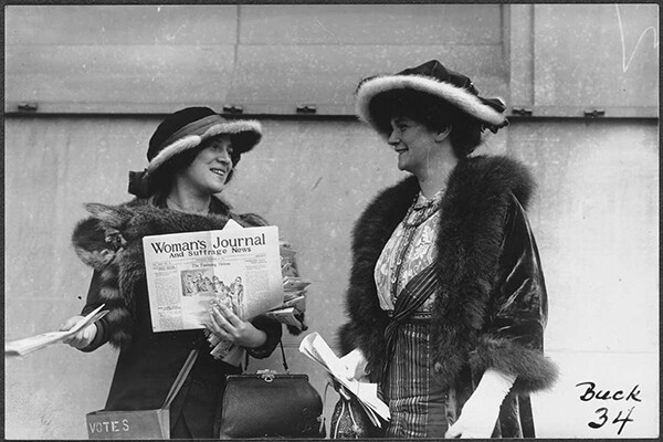 Two women in 1920 standing in fur lined coats and fancy hats, one holds a newspaper called Woman’s Journal and Suffrage News.