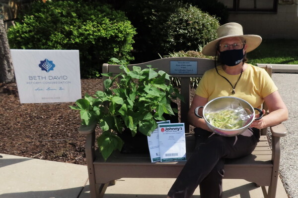 Person with a mask sitting on a bench holding a bowl of green beans next to a sign that says Beth David 