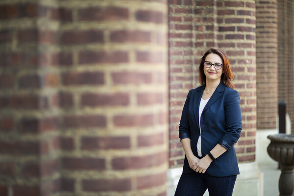 Smiling person standing arms held down, together and in front, outside of a brick building.  