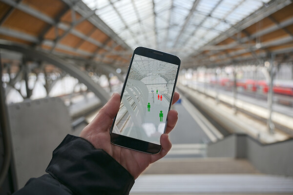 hand holding a smartphone in a public transit station tracking human images on the screen in red or green indicating covid exposure