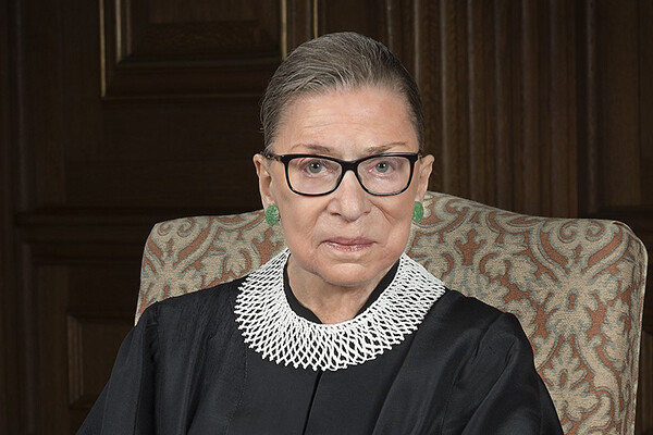 Person with hair pulled back, green earrings and black glasses looks into the camera, wearing Supreme Court robes and white lace collar.