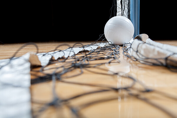 Volleyball on the ground on top of a collapsed net on a gym floor.