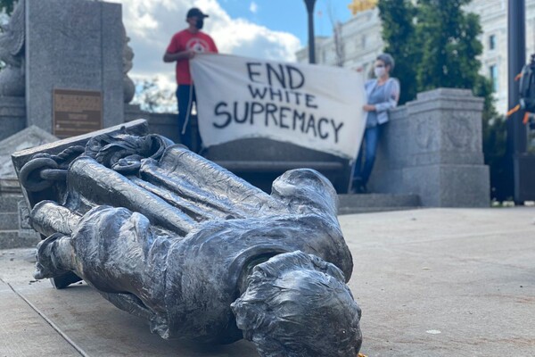A fallen statue of Columbus lays on the ground in the foreground, and two people hold up an End White Supremacy sign in the background.