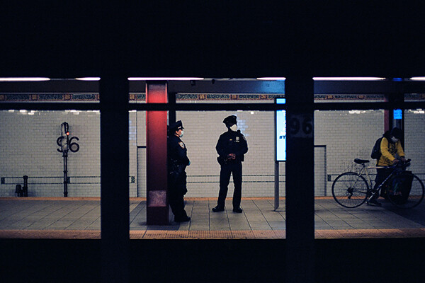 Two police officers in face masks standing in an underground subway stop, a person walking a bicycle walks past.